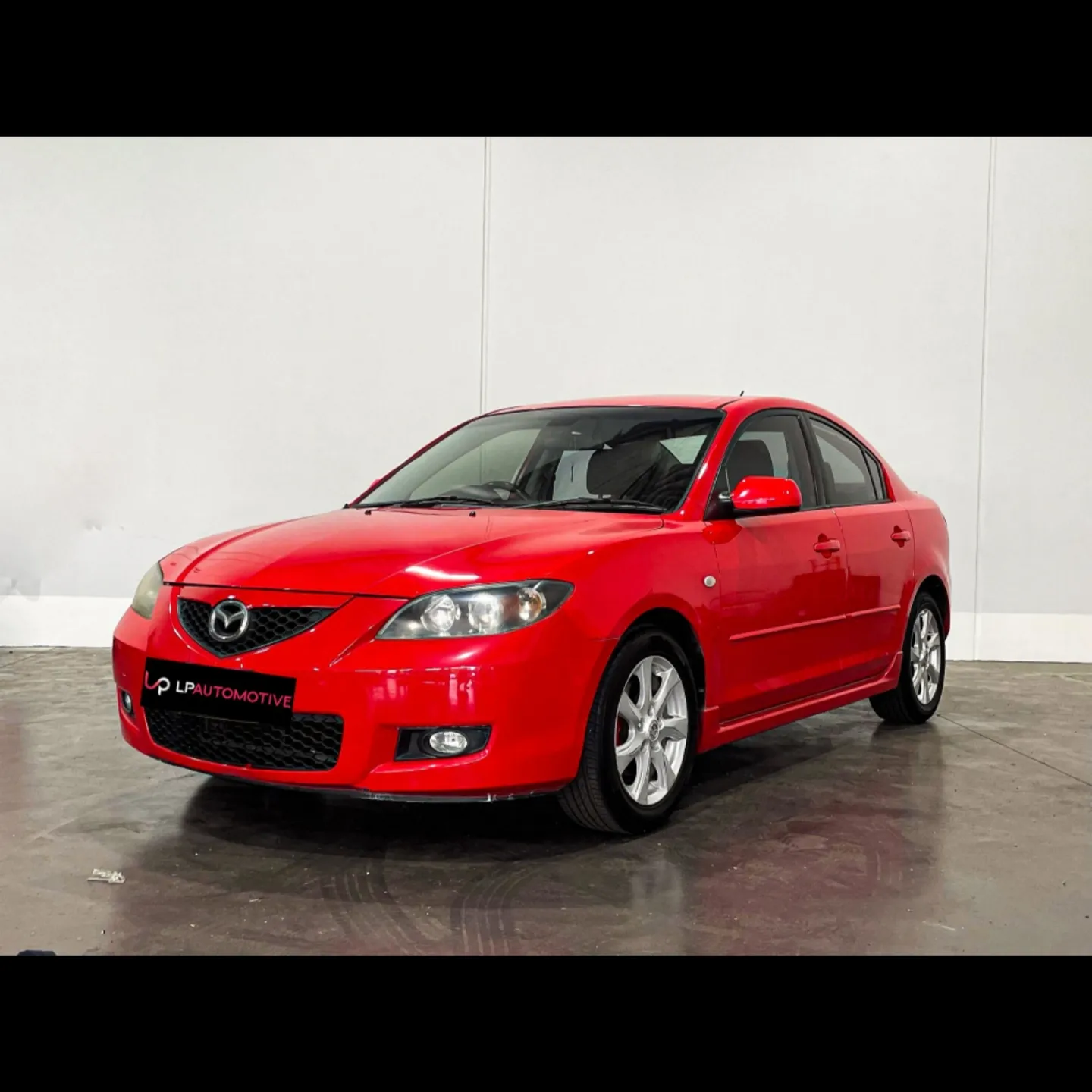 A red Mazda 3 sedan photographed against the white wall at LP Automotive, wearing an LP Automotive dealer plate